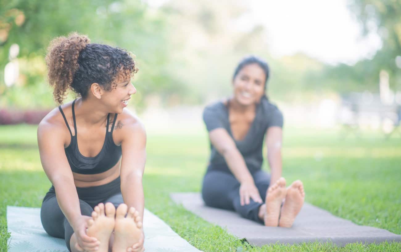 two woman touch their toes and do yoga on their pats in a park